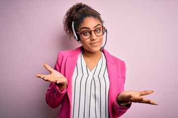 Young african american call center agent girl wearing glasses working using headset clueless and confused expression with arms and hands raised. Doubt concept.