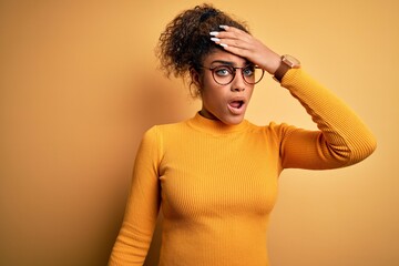 Young beautiful african american girl wearing sweater and glasses over yellow background surprised with hand on head for mistake, remember error. Forgot, bad memory concept.