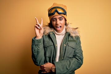 Young african american afro skier girl wearing snow sportswear and ski goggles smiling with happy face winking at the camera doing victory sign with fingers. Number two.