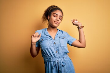 Young beautiful african american girl wearing denim dress standing over yellow background...
