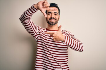 Naklejka premium Young handsome man with beard wearing casual striped t-shirt standing over white background smiling making frame with hands and fingers with happy face. Creativity and photography concept.