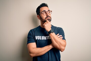 Handsome man with beard wearing t-shirt with volunteer message over white background with hand on chin thinking about question, pensive expression. Smiling with thoughtful face. Doubt concept.