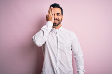 Young handsome man with beard wearing casual shirt standing over pink background covering one eye with hand, confident smile on face and surprise emotion.