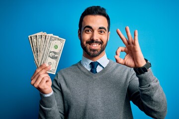 Young handsome man with beard holding bunch of dollars banknotes over blue background doing ok sign...