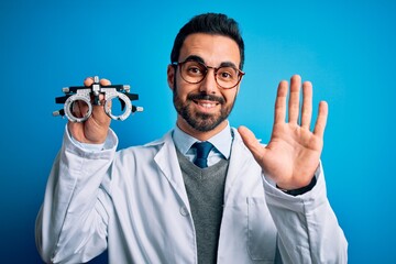 Young handsome optical man with beard holding optometry glasses over blue background Waiving saying hello happy and smiling, friendly welcome gesture