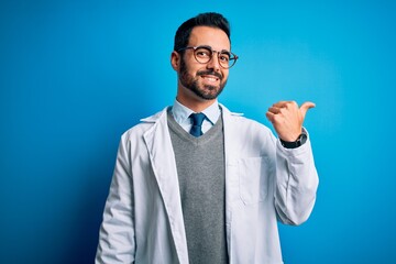 Young handsome doctor man with beard wearing coat and glasses over blue background smiling with happy face looking and pointing to the side with thumb up.