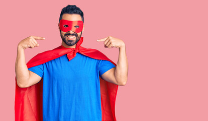 Young hispanic man wearing super hero costume looking confident with smile on face, pointing...