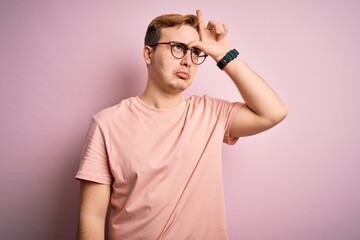 Young handsome redhead man wearing casual t-shirt standing over isolated pink background making fun of people with fingers on forehead doing loser gesture mocking and insulting.