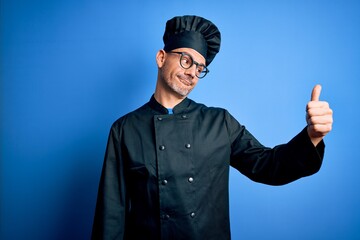 Young handsome chef man wearing cooker uniform and hat over isolated blue background Looking proud, smiling doing thumbs up gesture to the side