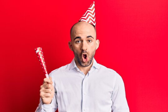 Young handsome bald man wearing party hat and trumpet scared and amazed with open mouth for surprise, disbelief face