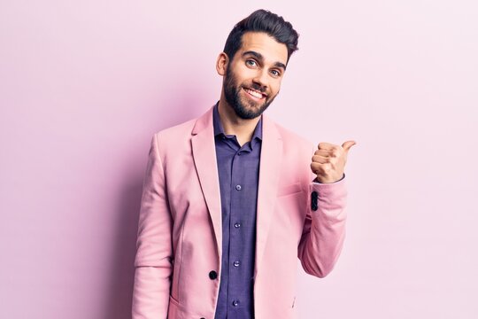 Young handsome man with beard wearing elegant jacket smiling with happy face looking and pointing to the side with thumb up.