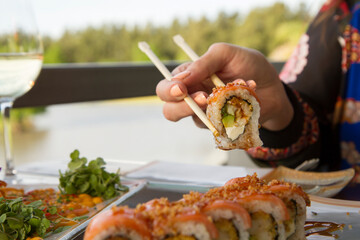 Gastronomy. Elegant sushi restaurant. Woman dining outside with a natural lake background. Young female hands holding a raw salmon sushi roll with the chopsticks.