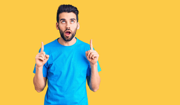 Young handsome man with beard wearing casual t-shirt amazed and surprised looking up and pointing with fingers and raised arms.