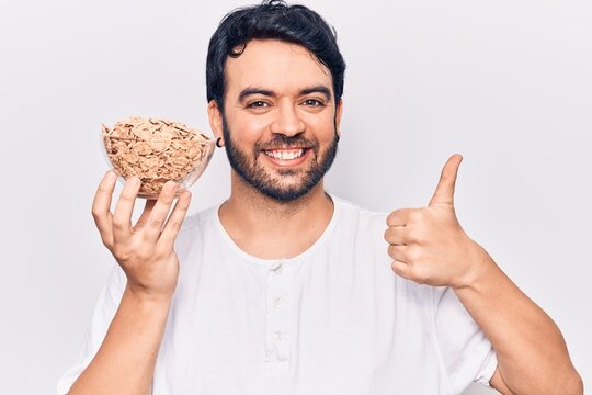 Young hispanic man holding cornflakes smiling happy and positive, thumb up doing excellent and approval sign