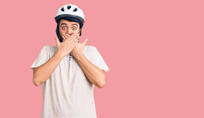 Young hispanic man wearing bike helmet shocked covering mouth with hands for mistake. secret concept.