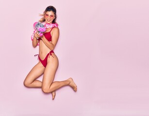 Fototapeta na wymiar Young beautiful girl on vacation wearing bikini and hawaiian lei smiling happy. Jumping with smile on face holding water gun over isolated pink background