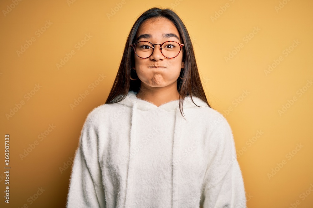 Sticker young beautiful asian woman wearing casual sweater and glasses over yellow background puffing cheeks