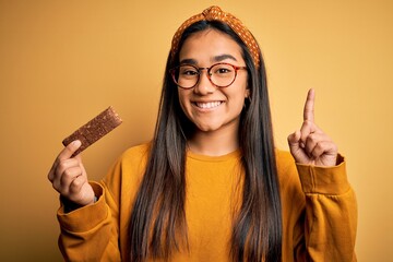 Young beautiful asian woman eating healthy protein bar over isolated yellow background surprised with an idea or question pointing finger with happy face, number one