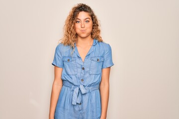 Young beautiful woman with blue eyes wearing casual denim dress over white background puffing...