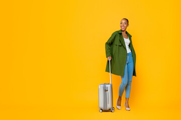 Full length portrait of travelling happy young African American tourist woman standing while holding luggage in isolated studio yellow background