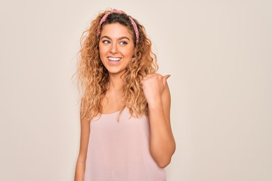 Young beautiful woman with blue eyes wearing casual t-shirt and diadem over pink background smiling with happy face looking and pointing to the side with thumb up.
