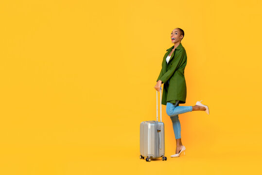 Full length portrait of excited young African American tourist woman holding luggage on one leg raised standing gesture ready to travel in isolated studio yellow background