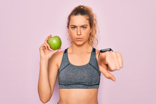 Young beautiful blonde sporty woman with blue eyes holding healthy green apple fruit pointing with finger to the camera and to you, hand sign, positive and confident gesture from the front