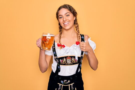 Beautiful blonde german woman with blue eyes wearing octoberfest dress drinking jar of beer happy with big smile doing ok sign, thumb up with fingers, excellent sign