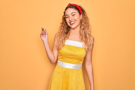 Beautiful blonde pin-up woman with blue eyes wearing diadem standing over yellow background with a big smile on face, pointing with hand finger to the side looking at the camera.
