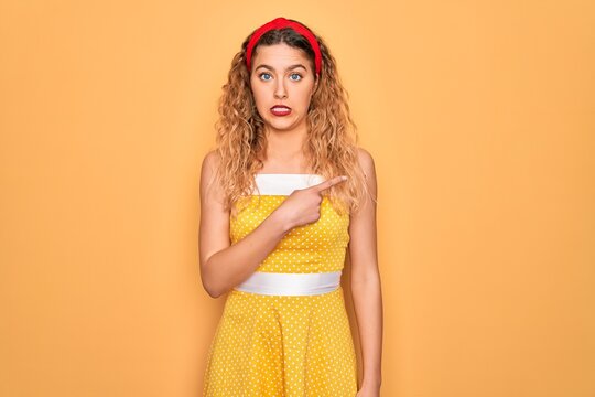 Beautiful blonde pin-up woman with blue eyes wearing diadem standing over yellow background Pointing aside worried and nervous with forefinger, concerned and surprised expression
