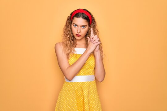 Beautiful blonde pin-up woman with blue eyes wearing diadem standing over yellow background Holding symbolic gun with hand gesture, playing killing shooting weapons, angry face