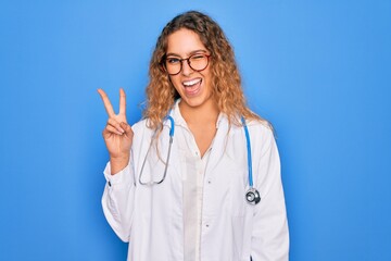 Young beautiful blonde doctor woman with blue eyes wearing coat and stethoscope smiling with happy face winking at the camera doing victory sign. Number two.