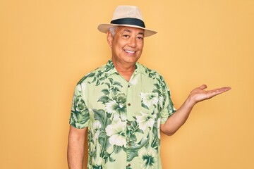 Middle age senior grey-haired man wearing summer hat and floral shirt on beach vacation smiling cheerful presenting and pointing with palm of hand looking at the camera.