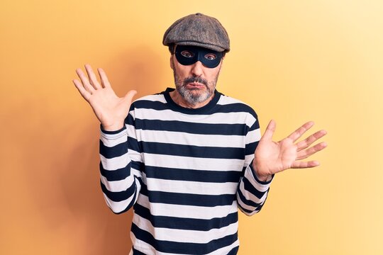 Middle age handsome burglar man wearing cap and mask over isolated yellow background clueless and confused with open arms, no idea and doubtful face.