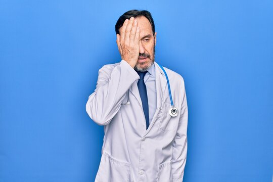 Middle age handsome doctor man wearing coat and stethoscope over isolated blue background surprised with hand on head for mistake, remember error. Forgot, bad memory concept.