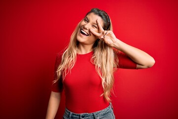 Young beautiful blonde woman wearing casual t-shirt standing over isolated red background Doing peace symbol with fingers over face, smiling cheerful showing victory
