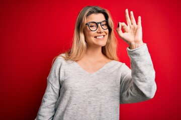 Young beautiful blonde woman wearing sweater and glasses over isolated red background smiling positive doing ok sign with hand and fingers. Successful expression.