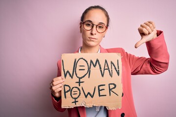 Young beautiful activist woman asking for women rights holding banner with power message with angry face, negative sign showing dislike with thumbs down, rejection concept