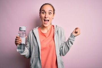 Young beautiful sporty woman doing sport drinking bottle with water to refreshment screaming proud and celebrating victory and success very excited, cheering emotion