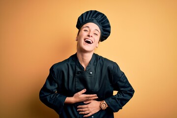 Young beautiful chef woman wearing cooker uniform and hat standing over yellow background smiling and laughing hard out loud because funny crazy joke with hands on body.