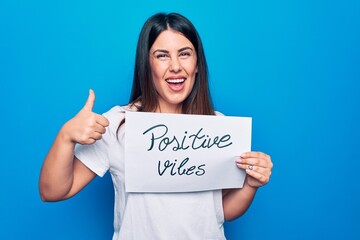 Young beautiful woman asking for optimist attitude holding paper with positive vibes message smiling happy and positive, thumb up doing excellent and approval sign