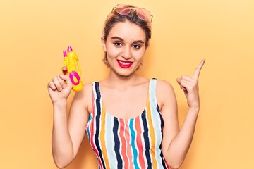 Young beautiful blonde woman wearing swimwear holding water gun smiling happy pointing with hand and finger to the side