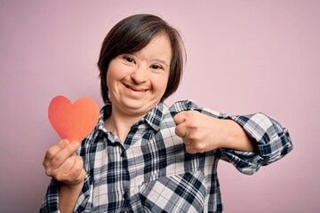 Young down syndrome romantic woman holding red heart paper shape over pink background screaming proud and celebrating victory and success very excited, cheering emotion