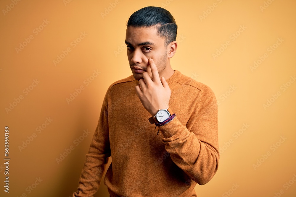 Wall mural Young brazilian man wearing casual sweater standing over isolated yellow background Pointing to the eye watching you gesture, suspicious expression