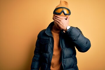 Young brazilian skier man wearing snow sportswear and ski goggles over yellow background covering eyes with hand, looking serious and sad. Sightless, hiding and rejection concept