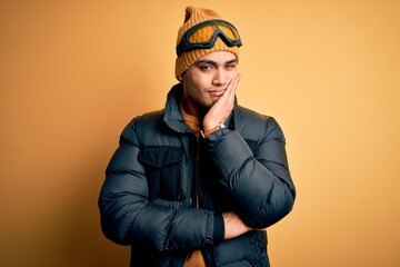 Young brazilian skier man wearing snow sportswear and ski goggles over yellow background thinking looking tired and bored with depression problems with crossed arms.