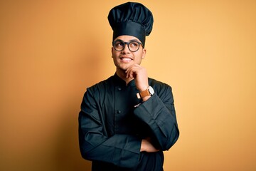 Young brazilian chef man wearing cooker uniform and hat over isolated yellow background with hand on chin thinking about question, pensive expression. Smiling and thoughtful face. Doubt concept.