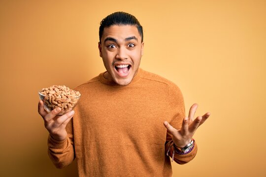 Young brazilian man holding bowl with healthy salty peanuts over isolated yellow background very happy and excited, winner expression celebrating victory screaming with big smile and raised hands