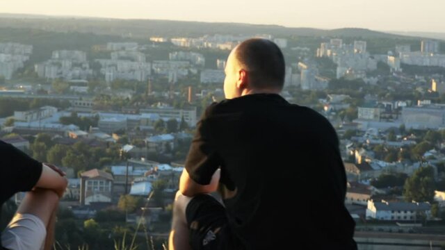 Two young caucasian men admiring modern cityscape sitting on cliff top together enjhoying each other company. Beautiful scenery.