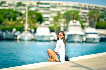 Fototapeta premium Cute smiling model girl in white shirt is sitting on pier near yachts.